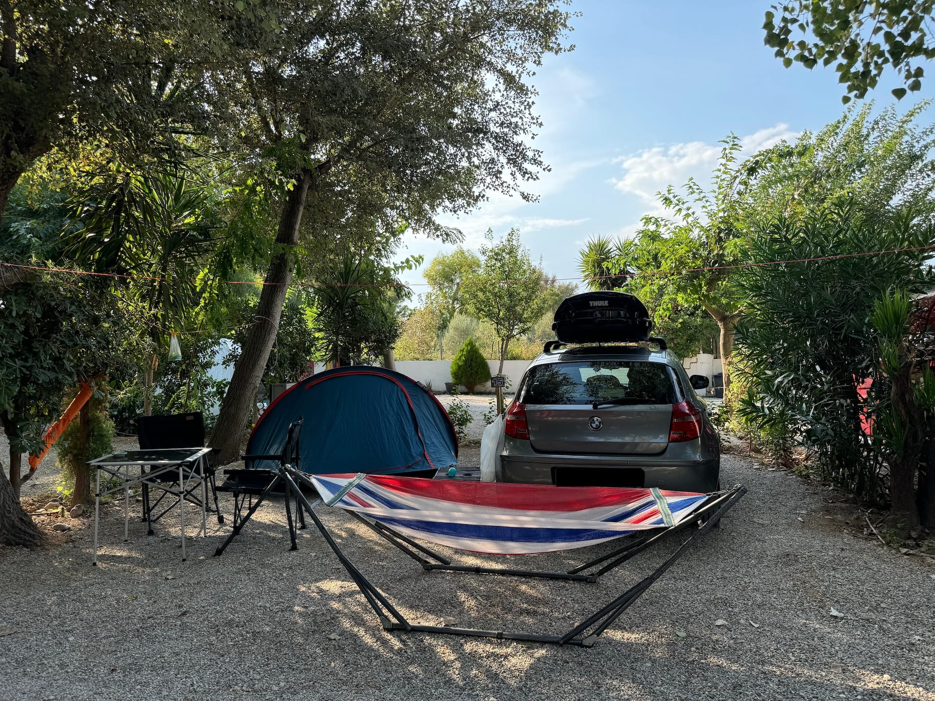 Kies-Stellplatz, umgeben von Pflanzen. Darauf Auto, Zelt, Tisch, zwei Stühle und eine Hängematte.