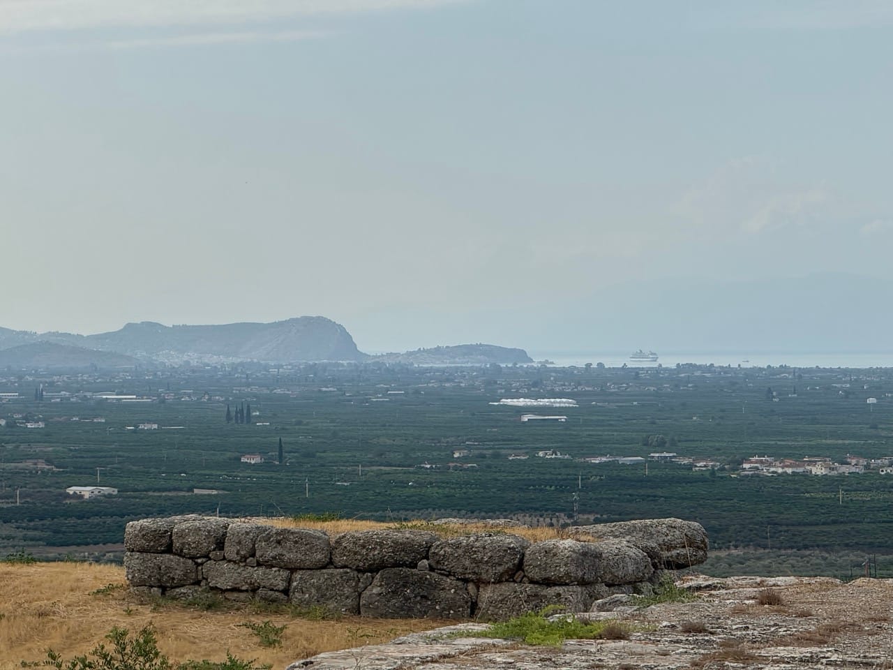 Im Vordergrund eine Ruine, im Hintergrund eine Ebene, ganz hinten Nafplio und das Meer.